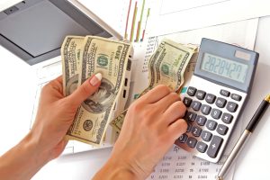 A person is using a calculator to count money while sitting at a desk with a laptop on it.