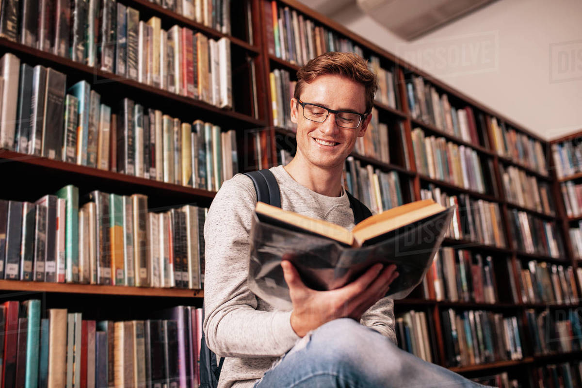 A young man with glasses is reading a book about investing while sitting in a library.
