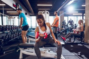 A woman in a white sports bra and black leggings is seated on a bench, lifting a dumbbell while two other people work out in the background.