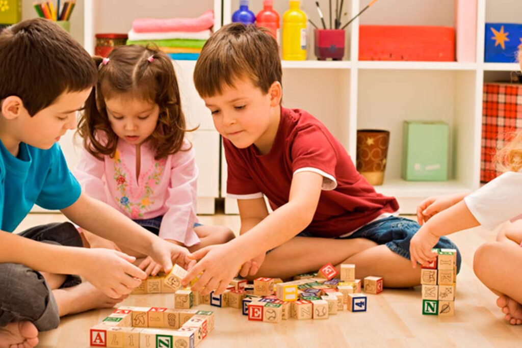 Three children are playing with wooden blocks on the floor, while another child looks on.