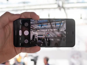 A hand holding a black smartphone in portrait orientation with the camera app open and pointed at a large room with people in it.