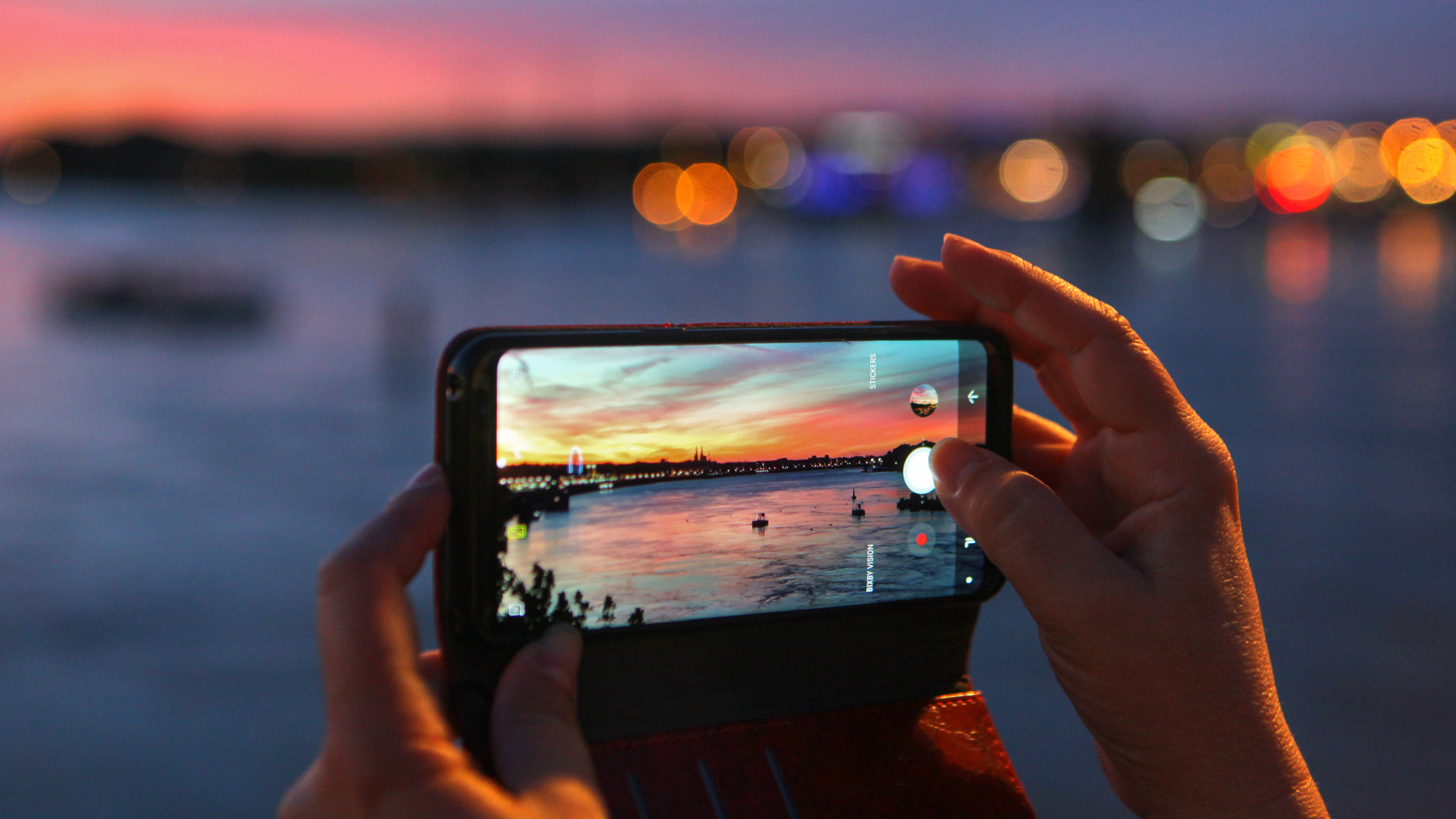A person is holding a smartphone in their hand and using the camera to take a picture of a sunset over a river. The smartphone is being held steady with both hands to prevent camera shake.