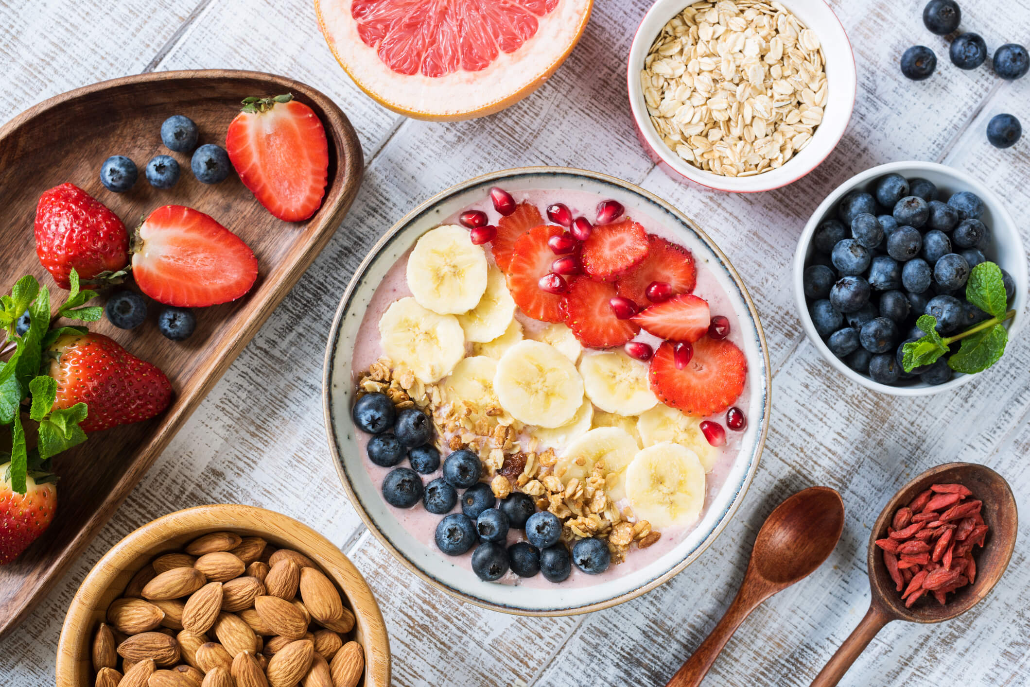 A variety of healthy breakfast options are arranged on a whitewashed wooden background including a bowl of smoothie topped with fruit and granola, a bowl of almonds, a bowl of blueberries, a bowl of strawberries, a bowl of oatmeal, a bowl of goji berries, and a sliced grapefruit.