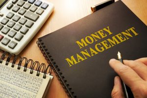 A person holding a pen in front of an open notebook with the words 'Money Management' on the cover, with a calculator and a notebook of financial calculations on the side.