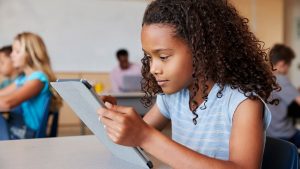 A young girl sits at her desk in a classroom, using a tablet to study with motivation.