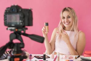 A young woman is sitting in front of a camera and pointing at a bottle of foundation while smiling.