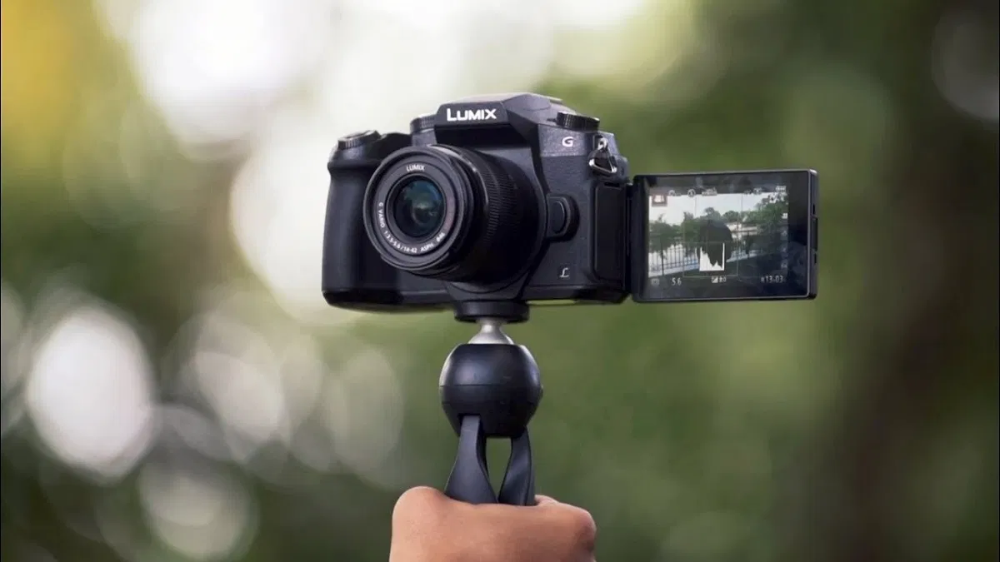 A small black camera with a flip-out screen, mounted on a tripod, for vlogging with high-resolution, high frame rate, interchangeable lenses, external microphone support, image stabilization, WiFi connectivity, Bluetooth connectivity, and a long battery life.