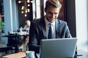 A man in a suit is sitting at a table in a cafe and using his laptop.