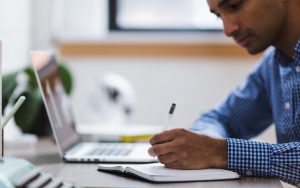 A man in a blue checkered shirt is writing in a notebook while sitting at a desk with an open laptop.