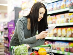 A woman leans on a shopping cart while looking at a grocery list with fresh produce in the cart.