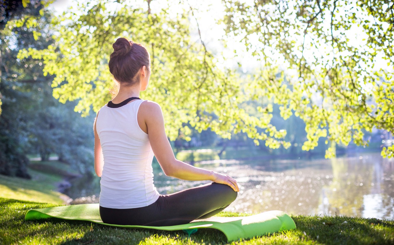 A person sits on a yoga mat in a park with their eyes closed and legs crossed in a peaceful meditation pose surrounded by trees and a lake.
