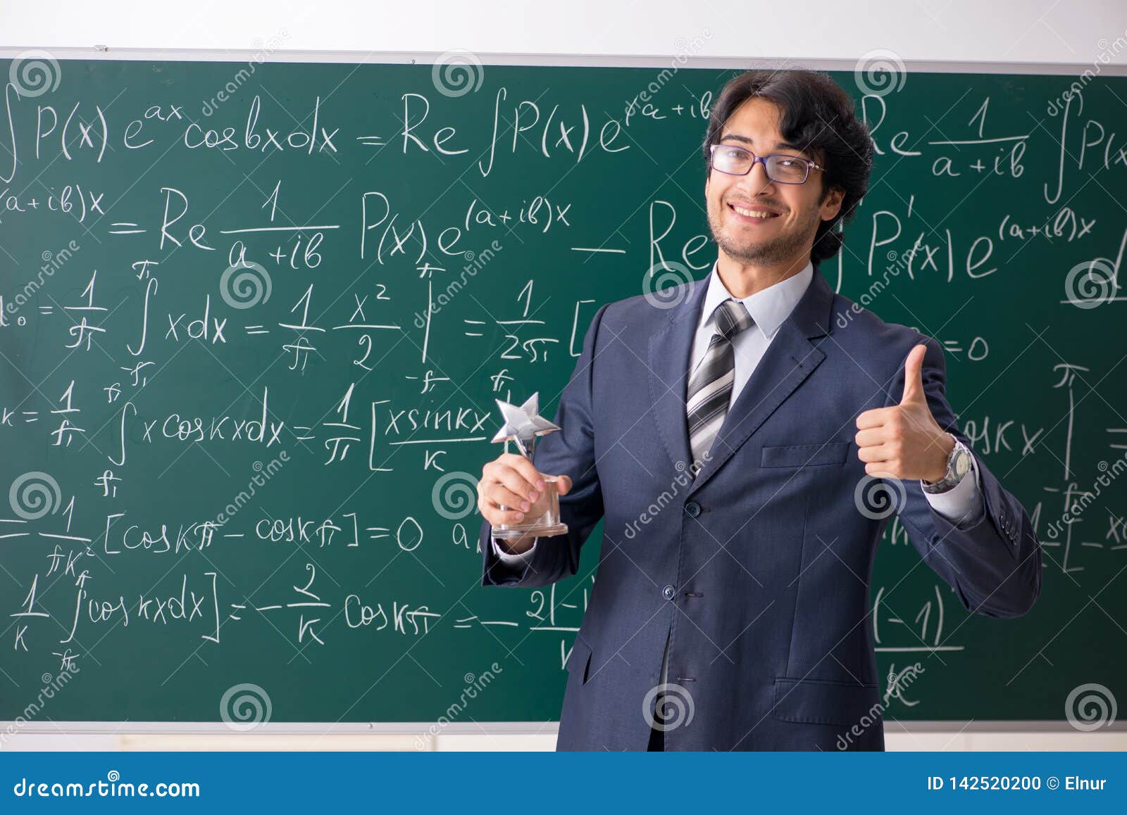 A math teacher standing in front of a blackboard full of math equations, holding a trophy, and giving a thumbs up.