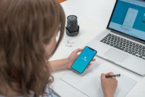 A woman is using a writing app on her phone while taking notes on a notepad with a pen.