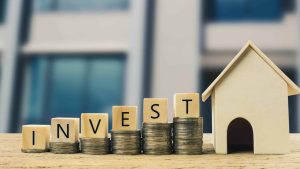 A wooden house sits next to a set of wooden blocks that spell out 'INVEST' sitting on top of stacks of coins.