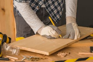 A person wearing protective gloves is using a pencil to mark a measurement on a piece of wood while working on a DIY technology project.