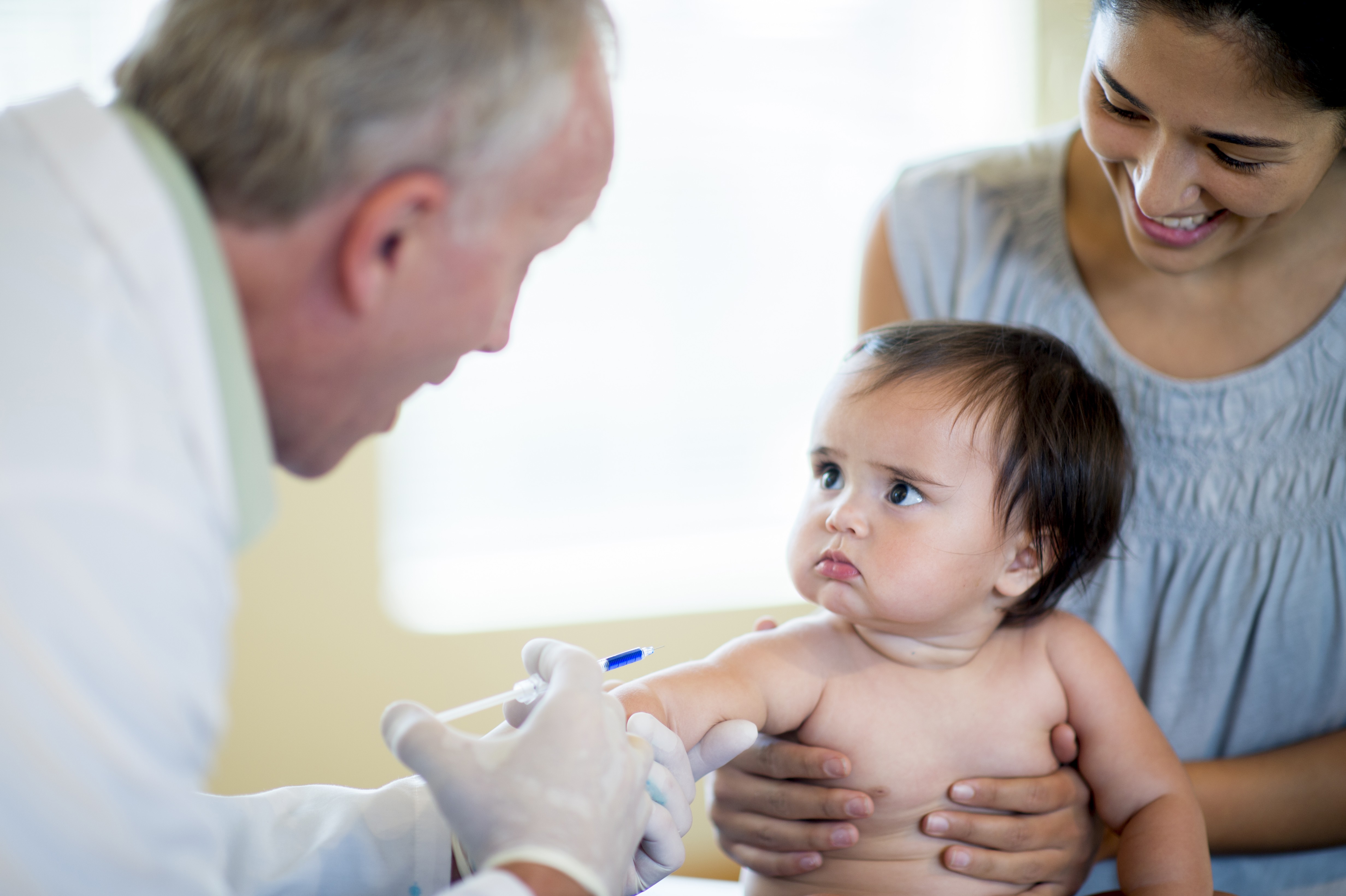 A smiling doctor in a white coat and gloves is administering a vaccination shot to a baby held by a smiling parent.