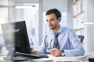 A man in a blue shirt and tie is sitting at a desk and looking at a computer monitor while holding his glasses.