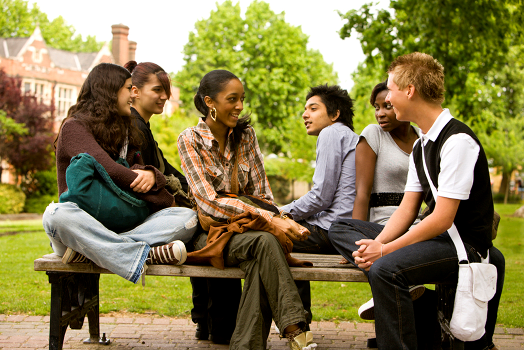 A group of diverse young people are sitting on a park bench and talking.