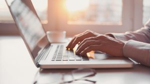 A person is typing a keyword research on a laptop in a brightly lit room.