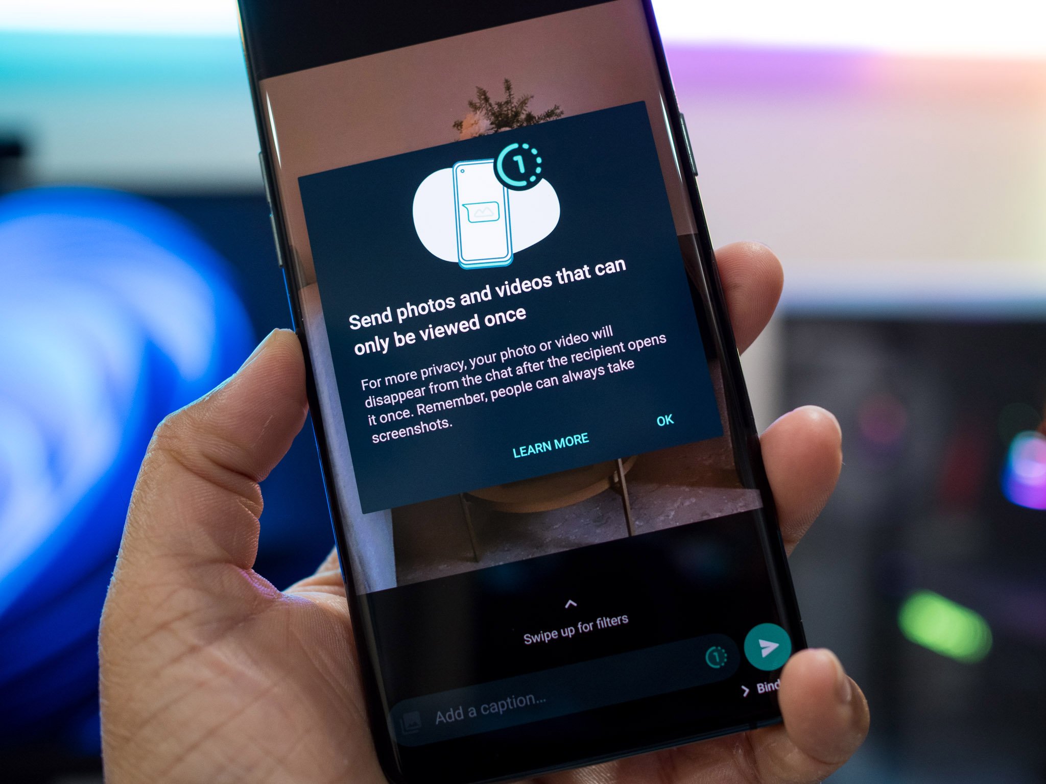 A hand holding a phone with a close up of the screen which shows a pop up with text 'Send photos and videos that can only be viewed once. For more privacy, your photo or video will disappear from the chat after the recipient opens it once. Remember, people can always take screenshots. Learn More. OK.'