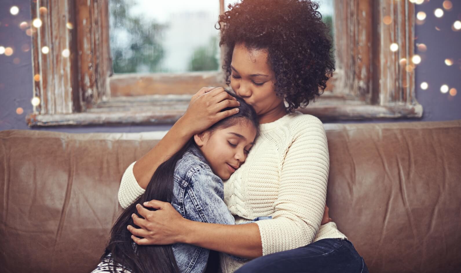 A mother and her daughter are sitting on a couch together. The mother is holding the daughter in her arms and the daughter is resting her head on the mother's shoulder. They are both smiling and look happy.