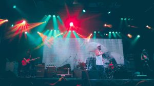 A brightly lit stage with a band performing in front of a large screen with a budget spreadsheet on it.