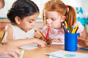 Two preschool girls are sitting at a table in a classroom. They are drawing and talking to each other.