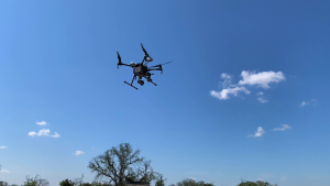 A black drone is flying in a blue sky with white clouds inspecting a wind turbine.