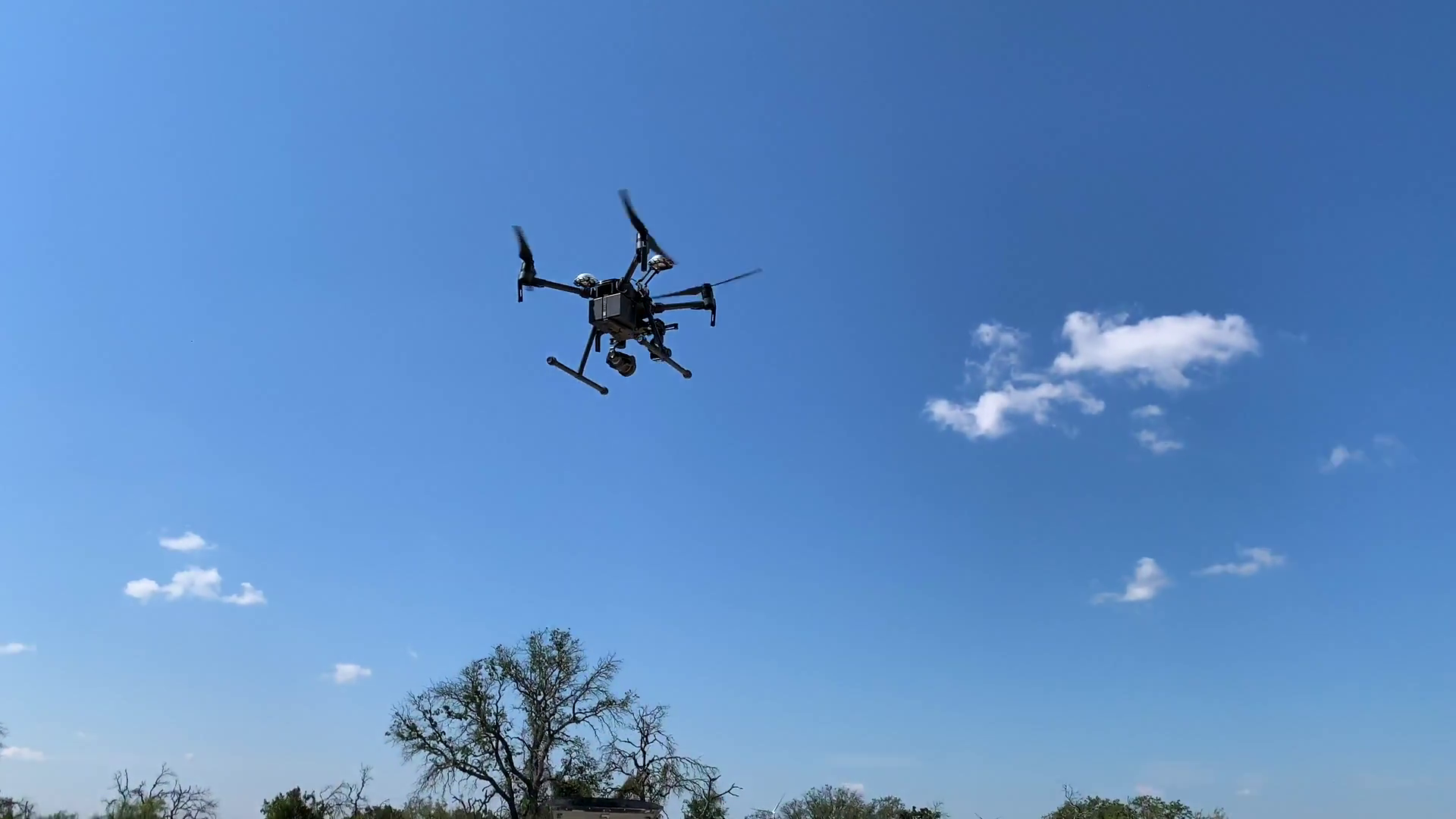 A black drone is flying in a blue sky with white clouds inspecting a wind turbine.