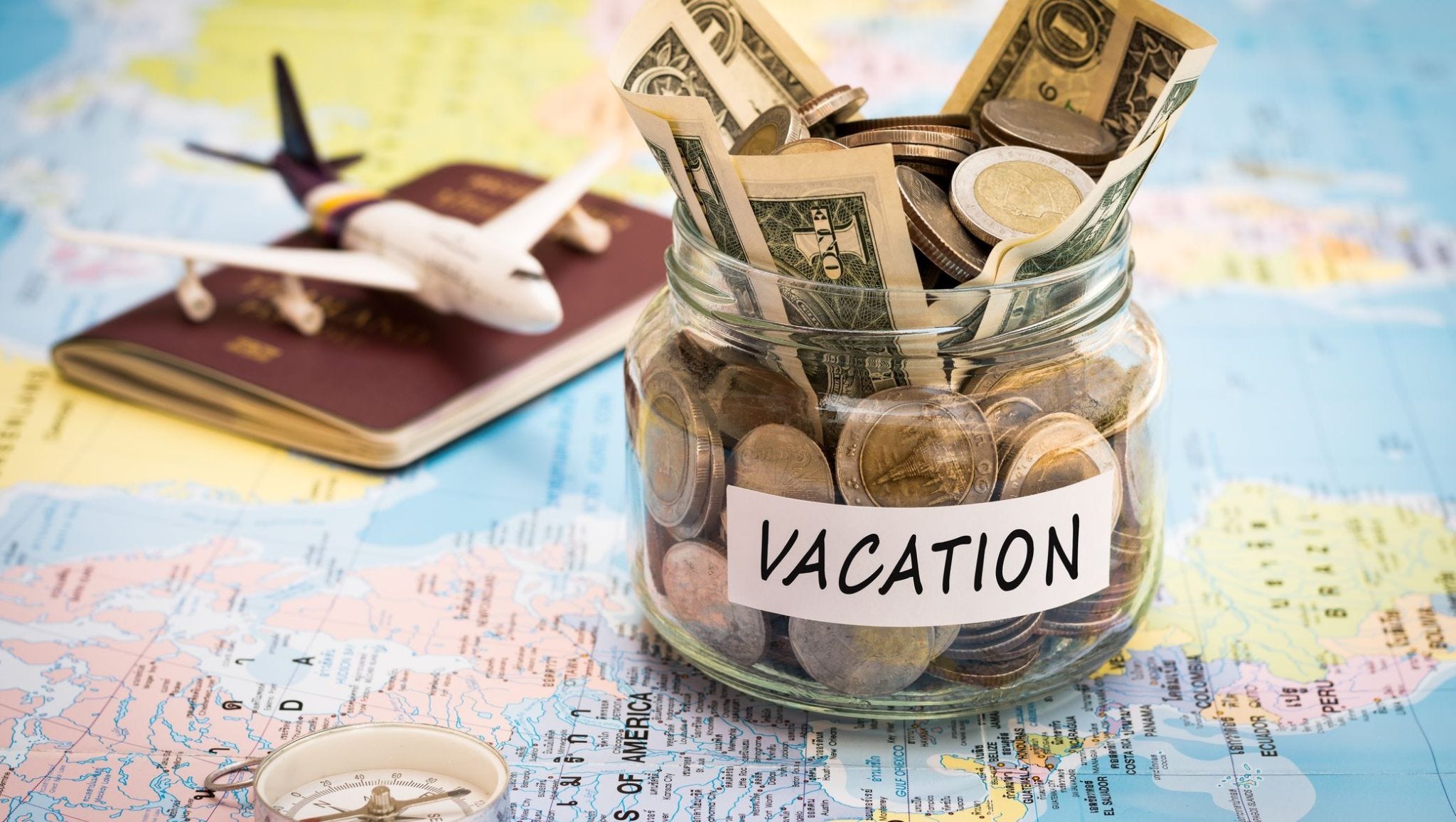 A jar filled with money and labeled 'VACATION' sits on top of a detailed world map with a toy airplane and a compass beside it.