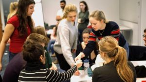 Students of diverse ethnicities engaged in active learning in a classroom with a whiteboard and colorful chairs.