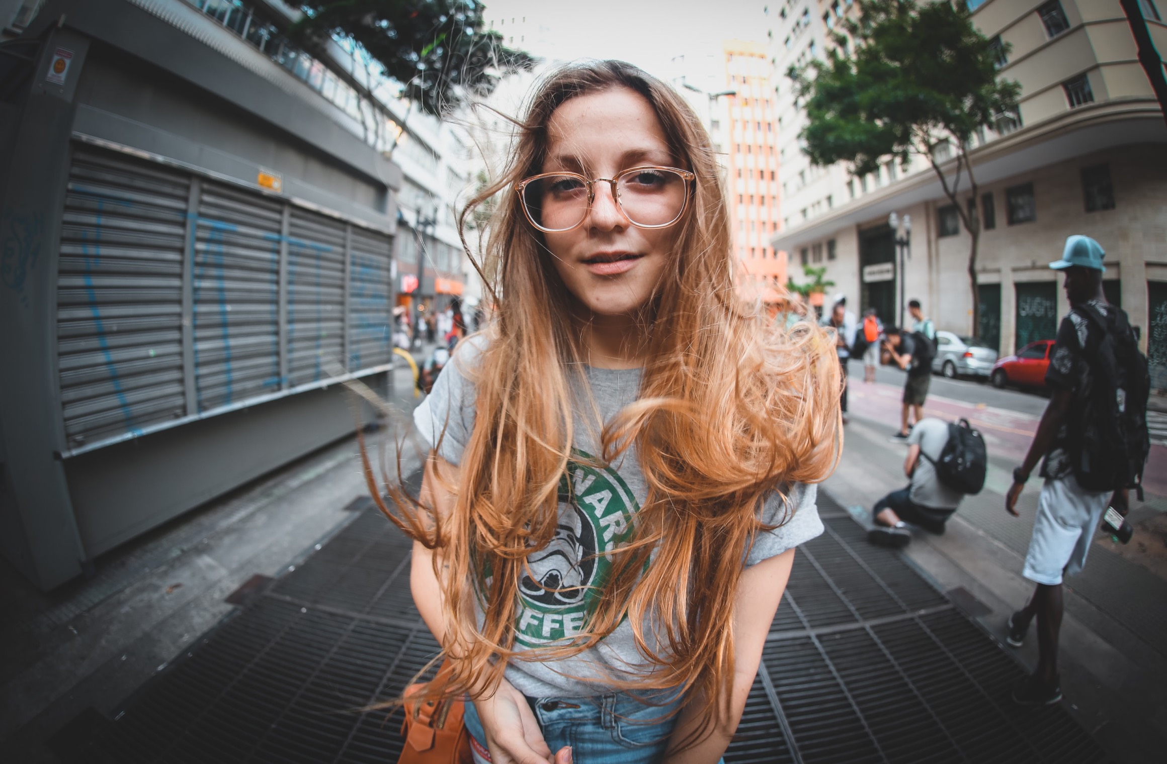 A young woman with long blond hair and glasses smiles at the camera on a busy city street with an ultra wide angle lens.