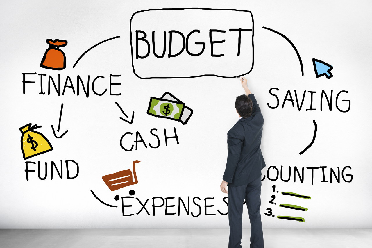 A businessman is drawing a budget plan on a whiteboard. The plan includes tips and tricks for budgeting for an event such as buying tickets, finding accommodation, and saving money on food and drinks.