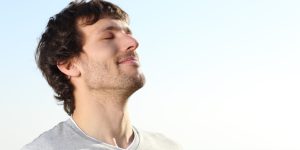 A man in a gray shirt is lying down with his eyes closed and taking slow deep breaths.