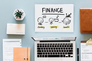 A desk with a laptop, notebook, cashbook, and a plant on it. The words 'funding', 'saving', and 'benefit' are written on a notepad, with a drawing of a smiling piggy bank and two smiley faces.