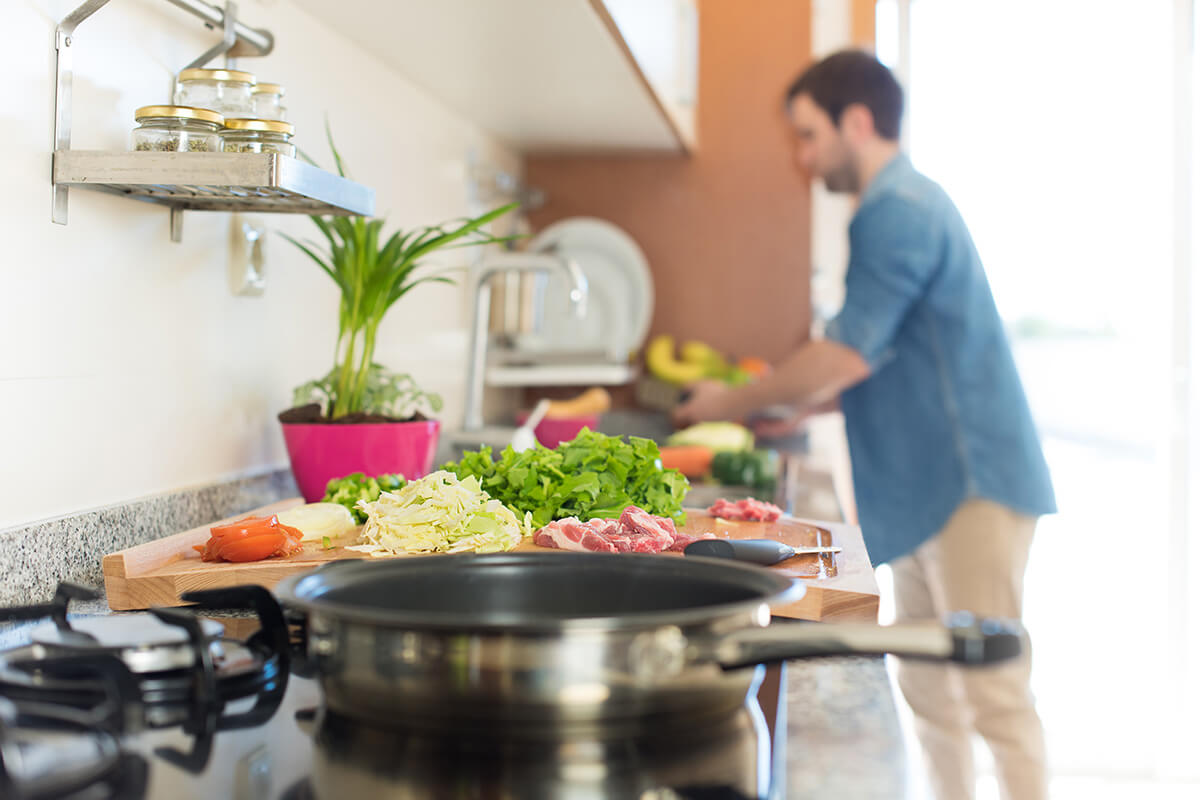 A person is chopping vegetables on a cutting board in the foreground with a pot sitting on the stove and a man is in the background blurry washing lettuce in a colander.