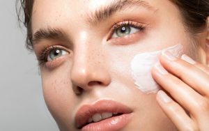 A young woman with freckles is applying a white cream to her face.