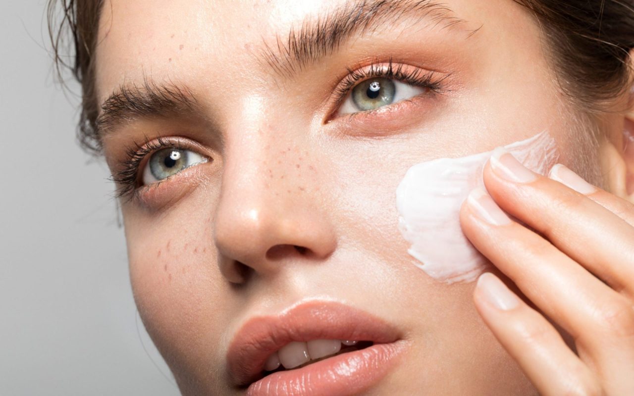 A young woman with freckles is applying a white cream to her face.