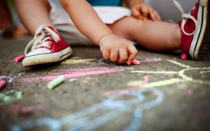 A toddler is drawing on the sidewalk with colorful chalk while sitting on the ground.