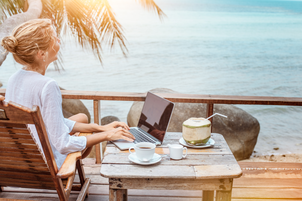 A person is sitting on a wooden deck overlooking the ocean, using a laptop to plan a vacation.