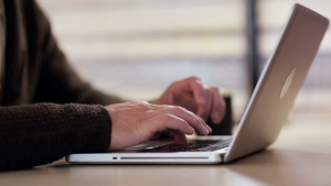 A person wearing a brown sweater types on an open laptop while sitting at a desk.