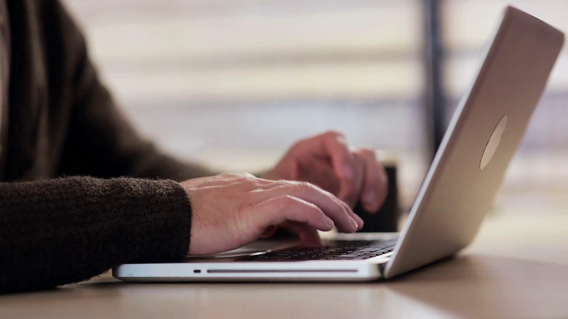 A person wearing a brown sweater types on an open laptop while sitting at a desk.