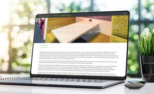 An image of a laptop computer on a wooden table with a plant beside it. The laptop is open and displaying a web page about global office design trends.