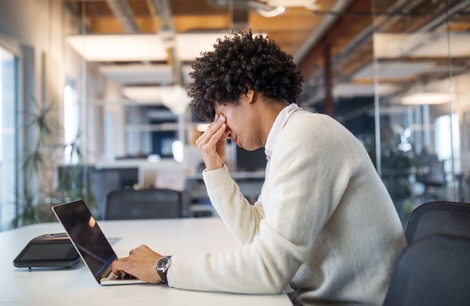 A person of color with curly hair wearing a white sweater works at a laptop and rubs their face, looking stressed.