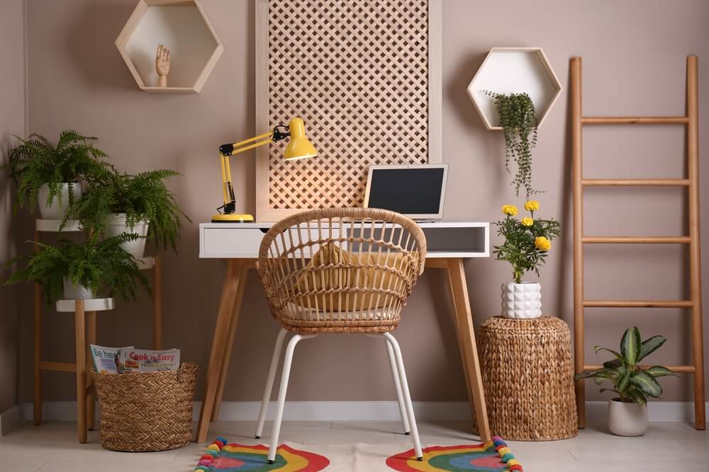 A cozy and comfortable study space for kids featuring a red velvet sofa, a round table, a yellow lamp, a rainbow rug, a wicker chair, a laptop, books, and plants.