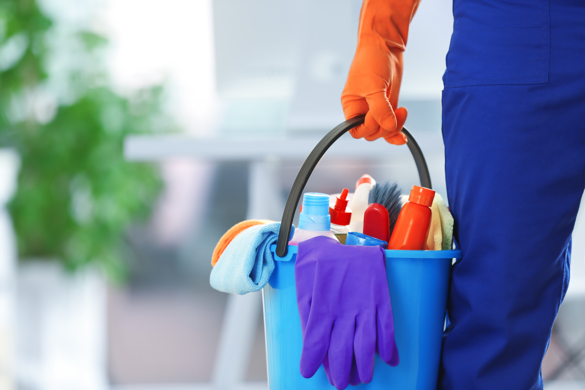 A person wearing blue overalls and orange gloves is holding a blue bucket filled with cleaning supplies. (illustrating tips for cleaning a house quickly and efficiently)
