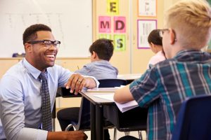 A teacher is talking to a student at his desk in a classroom, while other students look on.