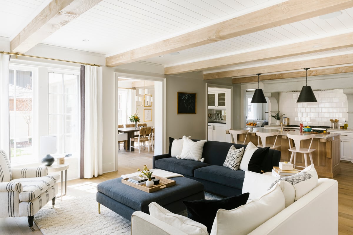 A living room with a large gray sectional, white walls, and wood beams on the ceiling furnished with a variety of cozy textiles, dramatic lighting, and personal accessories.