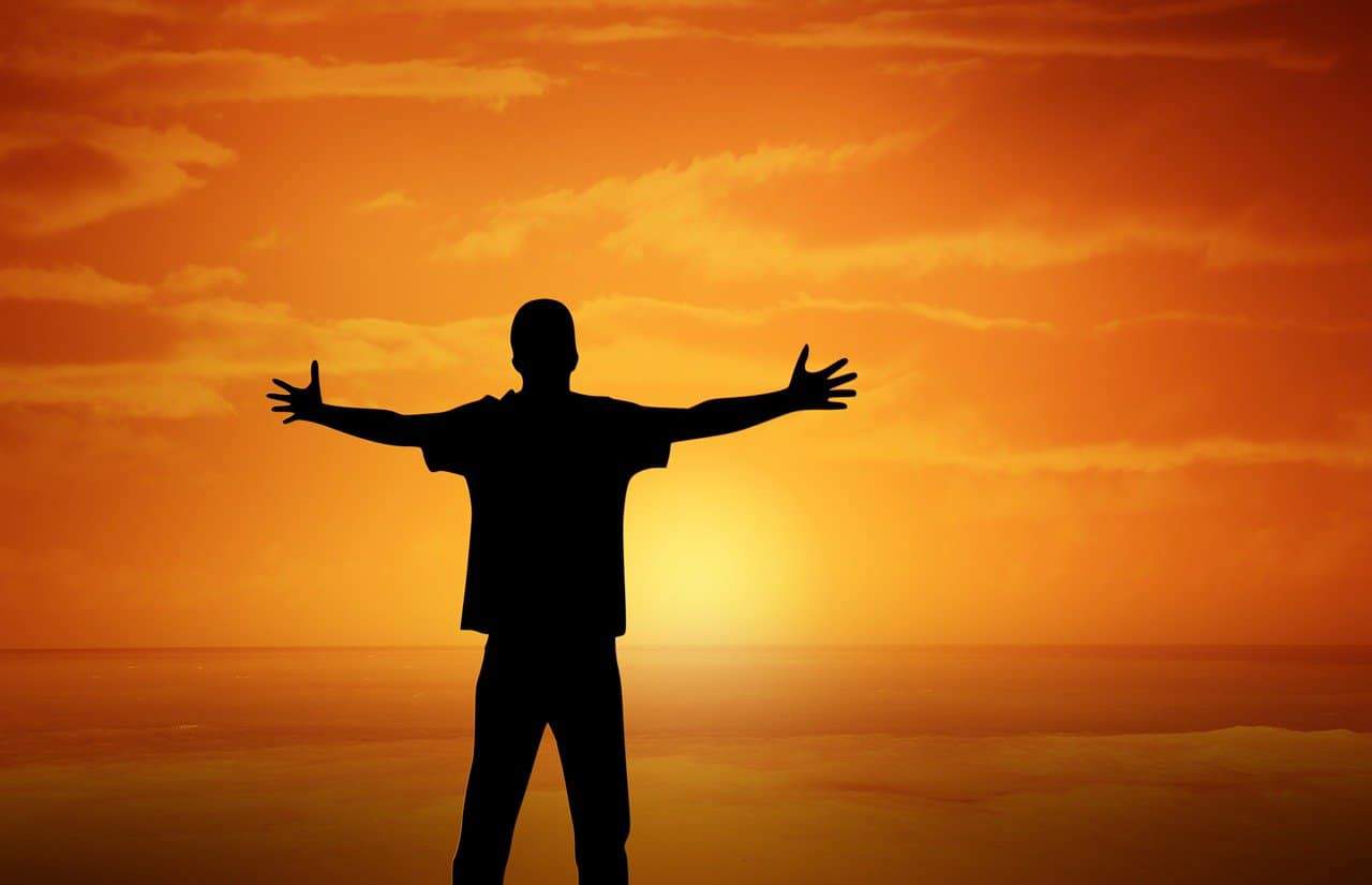 A person standing with their arms outstretched in front of a setting sun over water with the sky ablaze in orange.