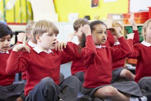 A group of children sit cross-legged on the floor in a classroom, singing and learning with songs.